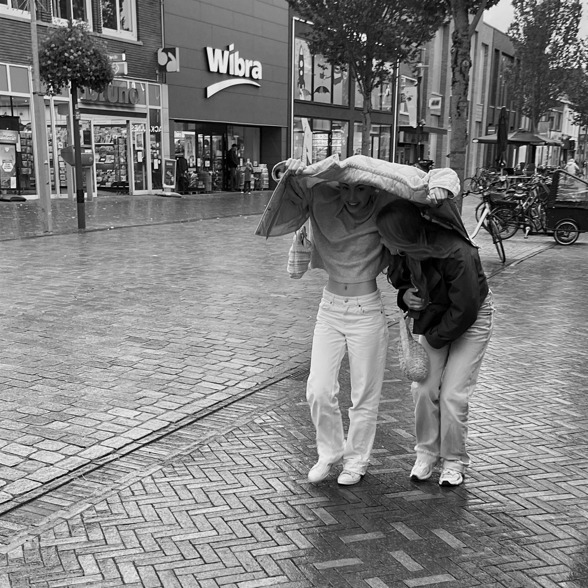 Two young women sheltering together beneath a coat in the rain.
